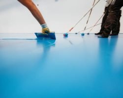 Workers applying a blue epoxy resin on a floor with rollers and spatulas in an industrial setting.