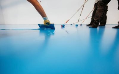 Workers applying a blue epoxy resin on a floor with rollers and spatulas in an industrial setting.